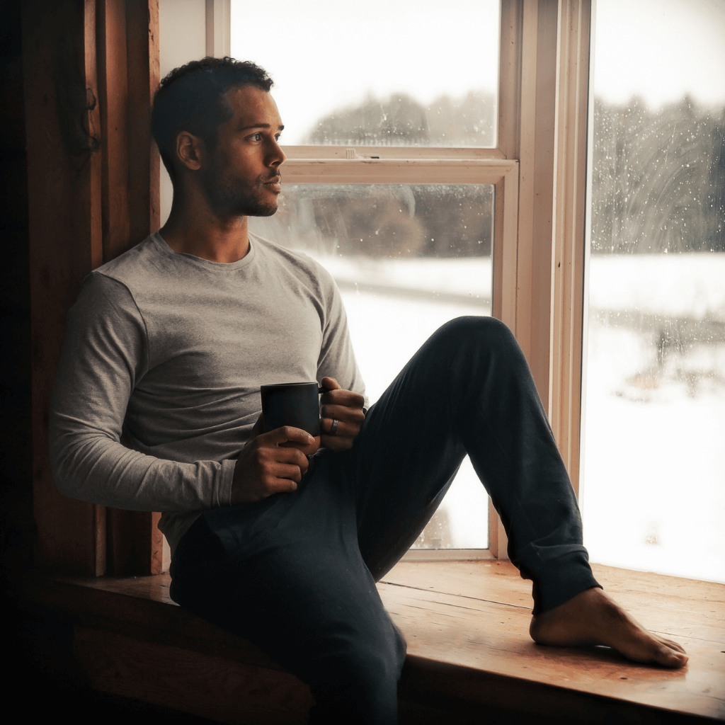 Relaxed man in cozy sweater sipping coffee by window, enjoying serene winter view.