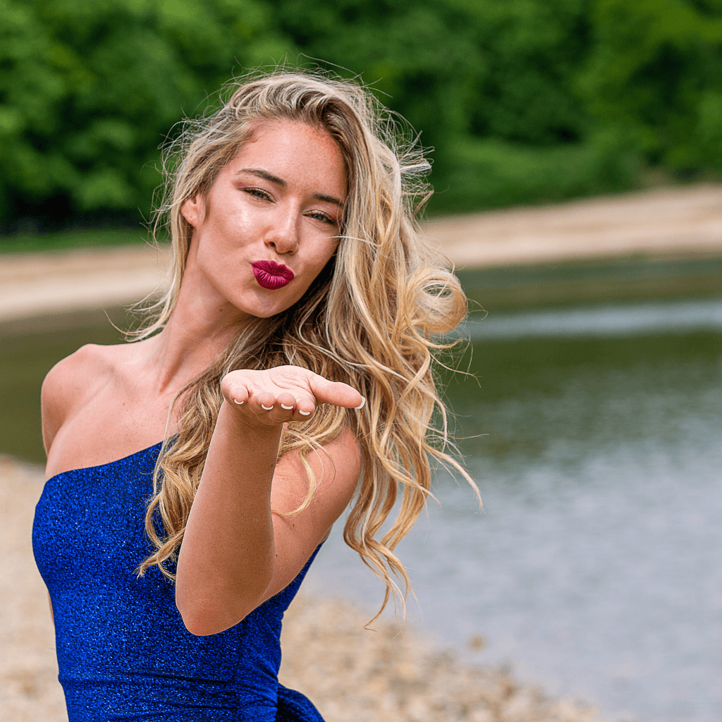 Beautiful woman blowing a kiss while wearing a blue dress near a serene lake setting.