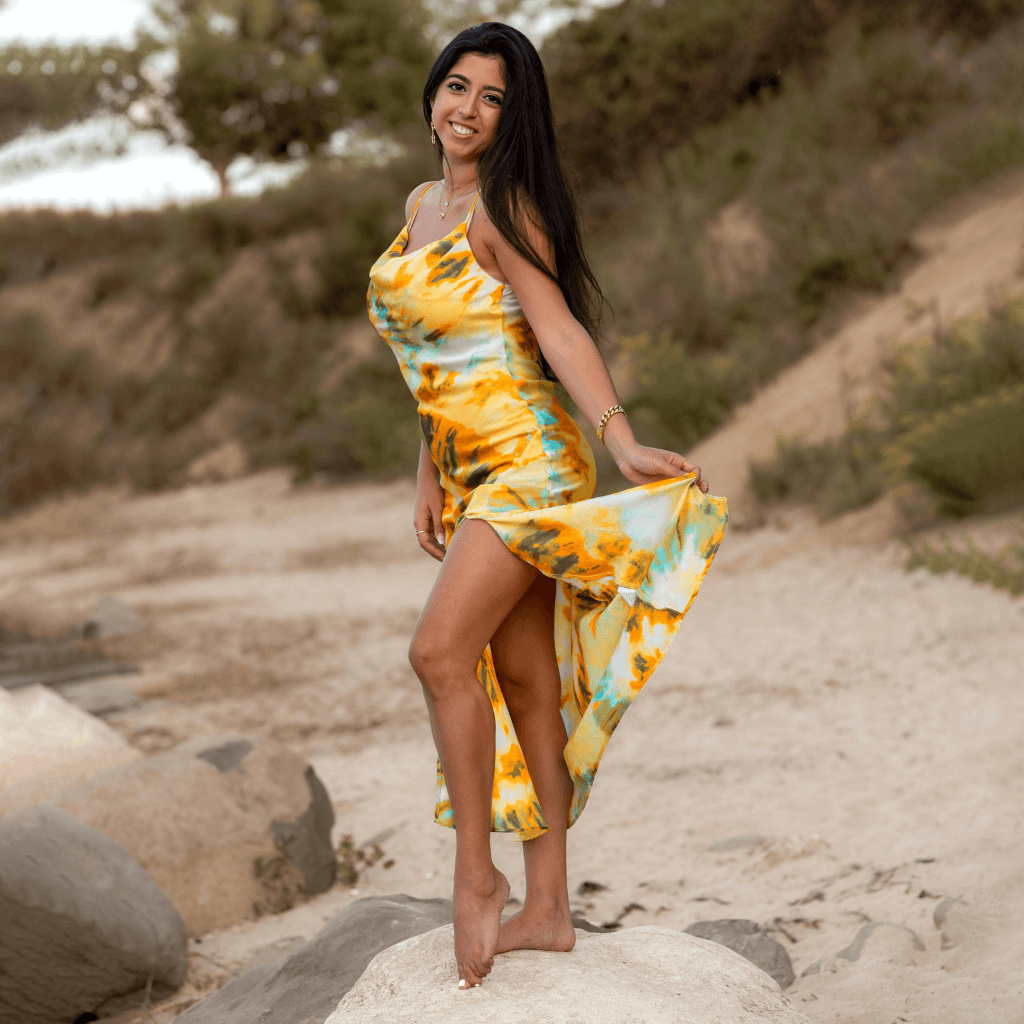Woman wearing a colorful dress with a high slit, posing playfully on a beach.