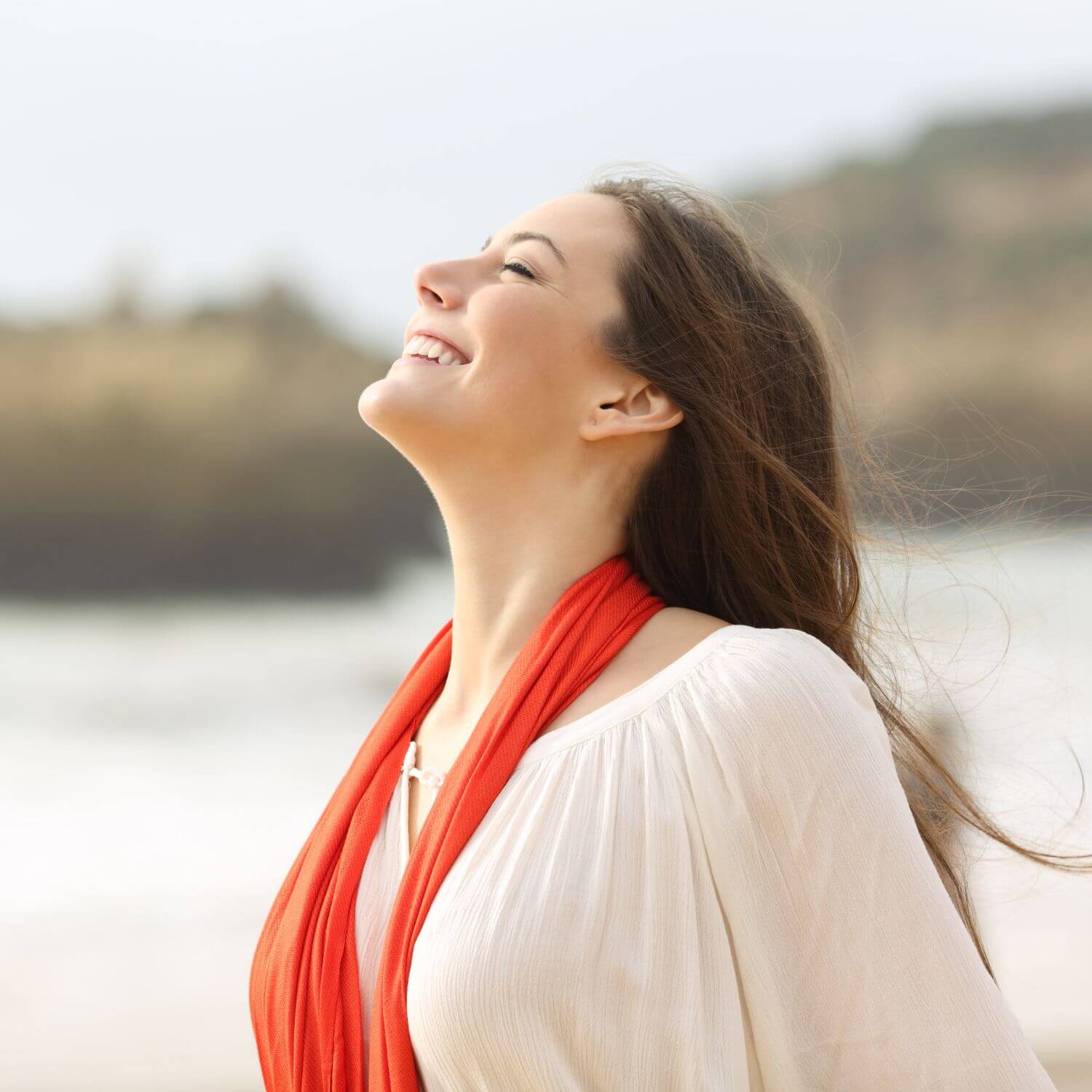 Smiling woman with flowing hair wearing a white top and red scarf at the beach, enjoying the fresh air.
