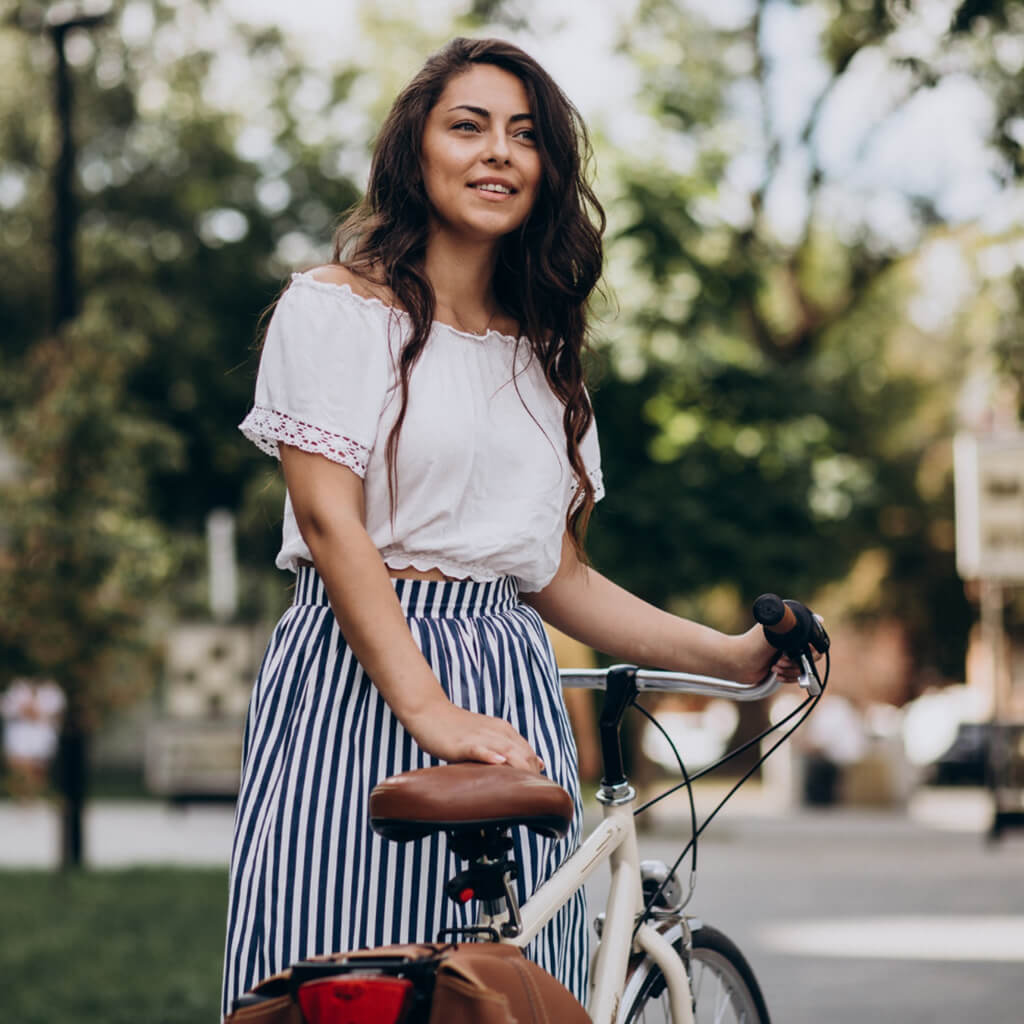 A stylish woman in a white top and striped skirt smiling while holding a bicycle outdoors.
